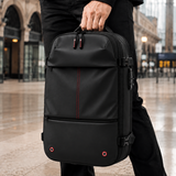 A person in black clothing holds a large black backpack-style travel bag with multiple zip compartments and buckle straps in an outdoor urban setting with blurred buildings in the background.
