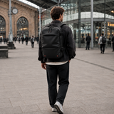 A man seen from behind walking through a modern train station, wearing a black backpack, dark jacket, and dark trousers.