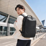 A man wearing a black backpack stands in front of a modern airport terminal building, looking back over his shoulder. The backpack is a matte black color with a clean, minimalist design.