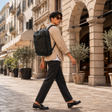 A man wearing a black backpack, beige shirt, black trousers, and black loafers walks along a cobblestone street in front of a stone building.