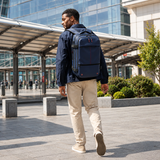 A man wearing a navy blue backpack with red accents walks outdoors in a modern urban setting. The backpack has multiple zippered compartments and appears to be made of durable nylon or polyester.