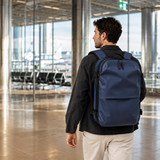A man wearing a navy blue backpack walks through a spacious, modern airport terminal with large windows and glass columns.