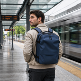 A man wearing a navy blue backpack stands on a train station platform with a train passing by in the background.