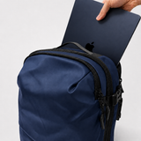 A navy blue backpack with zippers and hardware, shown with a hand inserting a dark blue laptop into its top pocket. The photo is shot against a clean, light studio background.