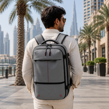 A man wearing a light beige shirt and trousers is shown from behind, carrying a gray laptop backpack with straps and accents in an outdoor city setting with palm trees and skyscrapers.