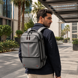 A man wearing a gray backpack with red accents, walking outdoors under a modern glass canopy with palm trees in the background.