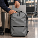 A gray backpack with zippers and handles, shown being held by a person whose hands and lower body are visible, with a silver laptop resting on top.