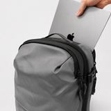 A gray backpack with zippers and trim, shown with a silver laptop being inserted into the top pocket. A hand is visible holding the laptop, and the Apple logo is visible on the laptop.