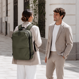 A man and a woman in beige suits are standing outdoors on a city sidewalk, conversing. The woman is wearing a dark olive green backpack, which is the product being showcased in this lifestyle setting.