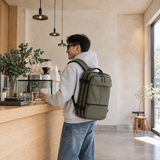 A man in a light hoodie and jeans stands at a cafe counter, wearing a green professional backpack with straps and zippers. The cafe has a warm, minimalist interior with hanging pendant lights and a wooden counter.
