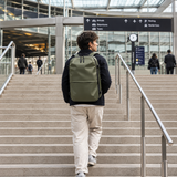 A man wearing a dark navy jacket, beige pants, and a green backpack walks up a large indoor staircase in a modern transit station.