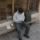 A man sits on a wooden bench in front of a shop window, wearing a hoodie and dark jeans while holding a camera. An olive green backpack rests on the bench beside him, along with a book and a small cup.