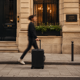 A person walking briskly on a city sidewalk while pulling a black hard-shell rolling suitcase in front of a beige stone building facade.
