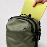 An olive green backpack with zippers and trim, shown with a hand inserting a yellow-green laptop into its main compartment against a light background.