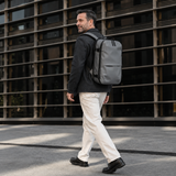 A man walking outdoors wearing a dark gray backpack, dressed in a blazer, white trousers, and shoes, set against a modern building with a dark grid pattern.