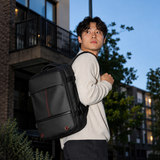 A man wearing a cream ribbed sweater and dark pants carries a black backpack with red accents while looking back over his shoulder in an outdoor urban setting at dusk.