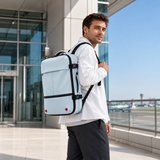A man wearing a white shirt and dark trousers walks outdoors carrying a light blue rolling suitcase backpack with straps and trim.