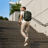 A man wearing a white t-shirt, beige trousers, and white sneakers walks up outdoor concrete stairs while carrying a dark green backpack.
