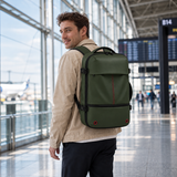 A man wearing a beige jacket and dark pants carries an olive green backpack with straps and trim in a modern airport terminal.