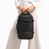 A woman wearing a white t-shirt and brown pants holds a black backpack against a paneled wall.